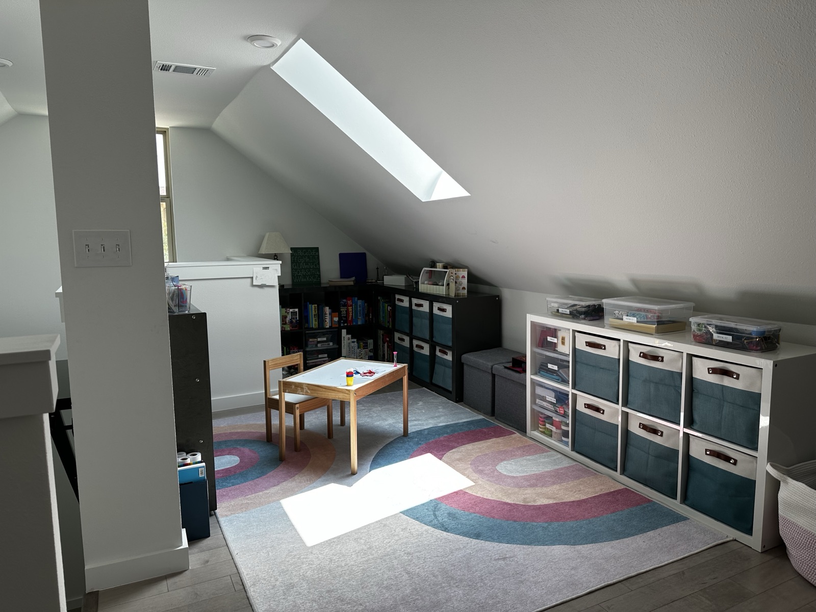 Attic playroom under a skylight with a child-size table, rainbow rug and fabric bins in built-in cubbies