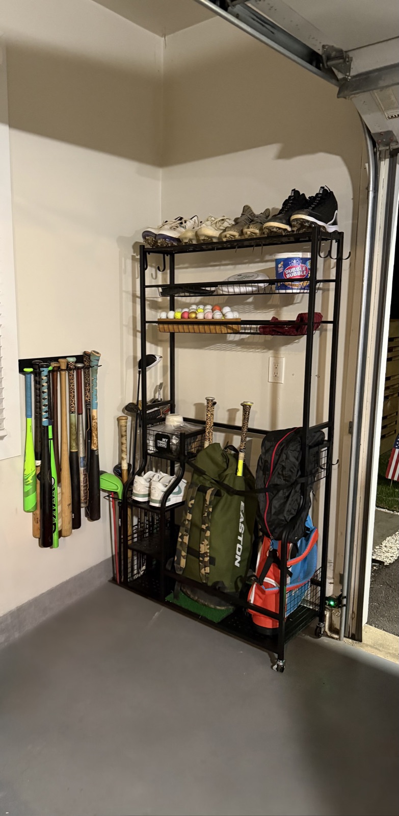 Garage corner with baseball bats racked on the wall and a rolling shelf holding cleats, balls and team bags, in Austin, TX