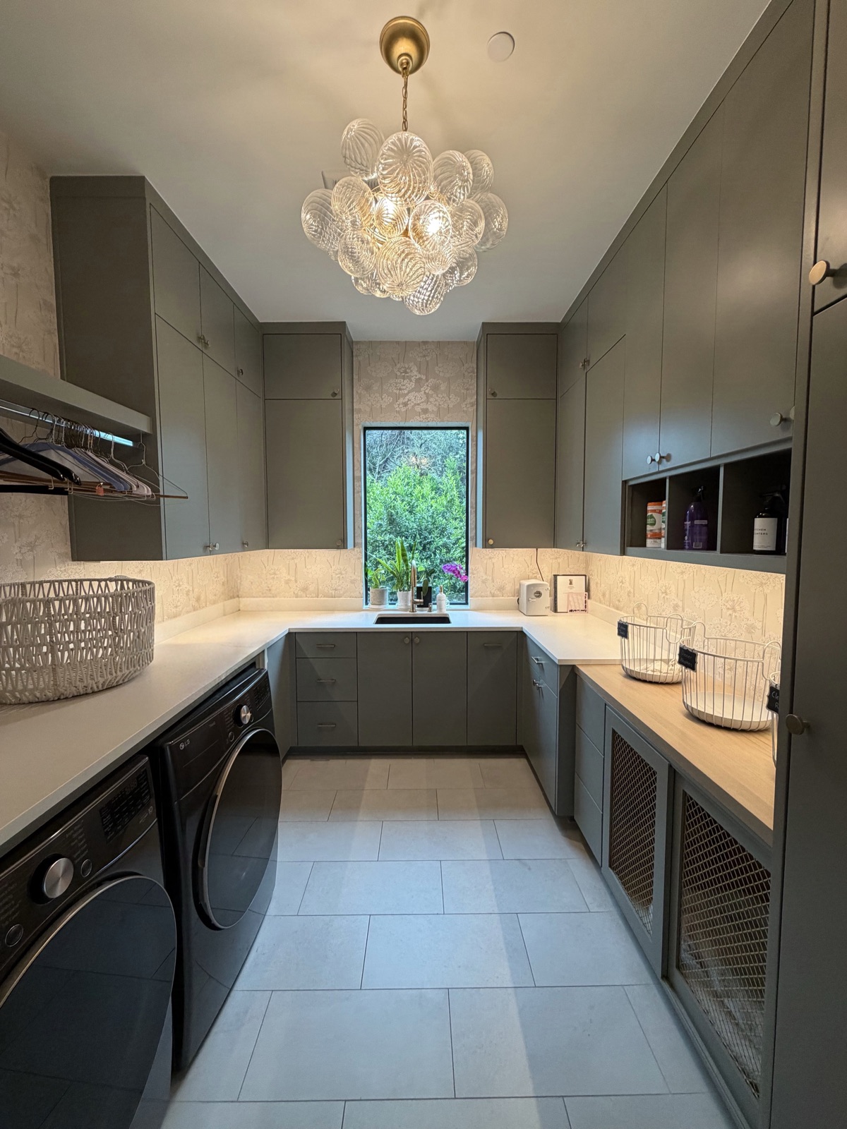 Laundry room with sage green cabinetry, white counters, front-loading washer and dryer and a glass bubble chandelier, in Austin, TX