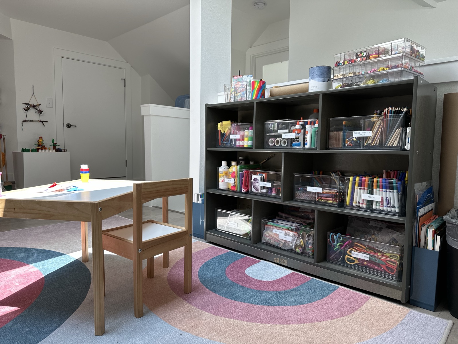 Playroom corner with a small wooden table and a tall black shelf of labeled clear bins holding craft supplies