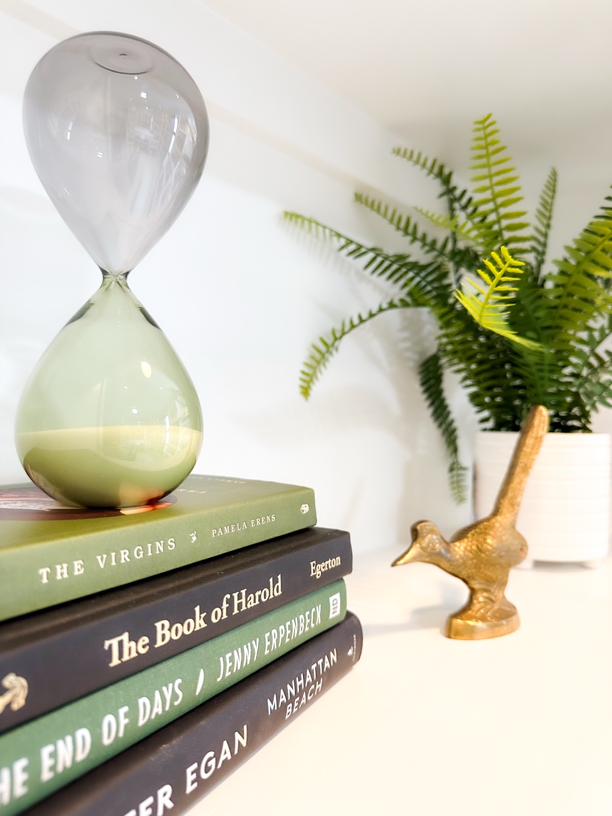 Hourglass on books, gold bird statue and green fern in a white square pot
